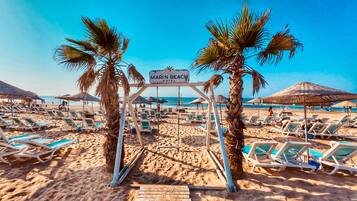 Una playa con sillas de descanso, palmeras y un cartel que indica 'Marin Beach Hotel'.