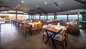 Un restaurante espacioso con varias mesas y sillas, amplias ventanas que ofrecen vista al mar y un diseño de techo moderno.