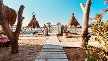Uma trilha na praia que leva a uma área arenosa com guarda-sóis de palha e espreguiçadeiras.