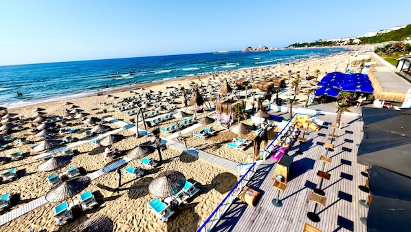 Una playa con numerosas tumbonas y sombrillas, una terraza de madera y vistas al mar.