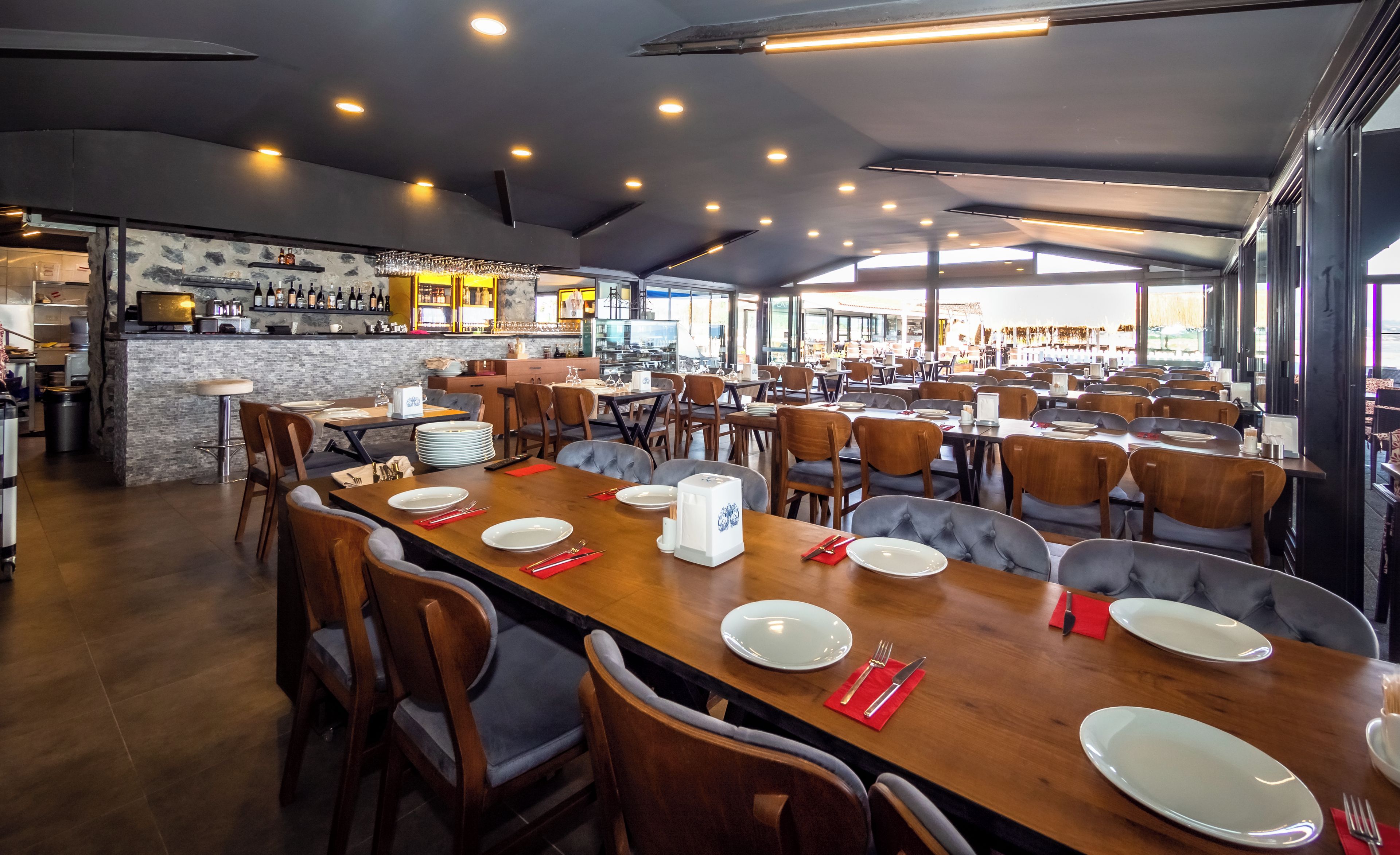 A spacious restaurant with multiple dining tables and chairs, a bar area with a wine display, and a view of the street outside.