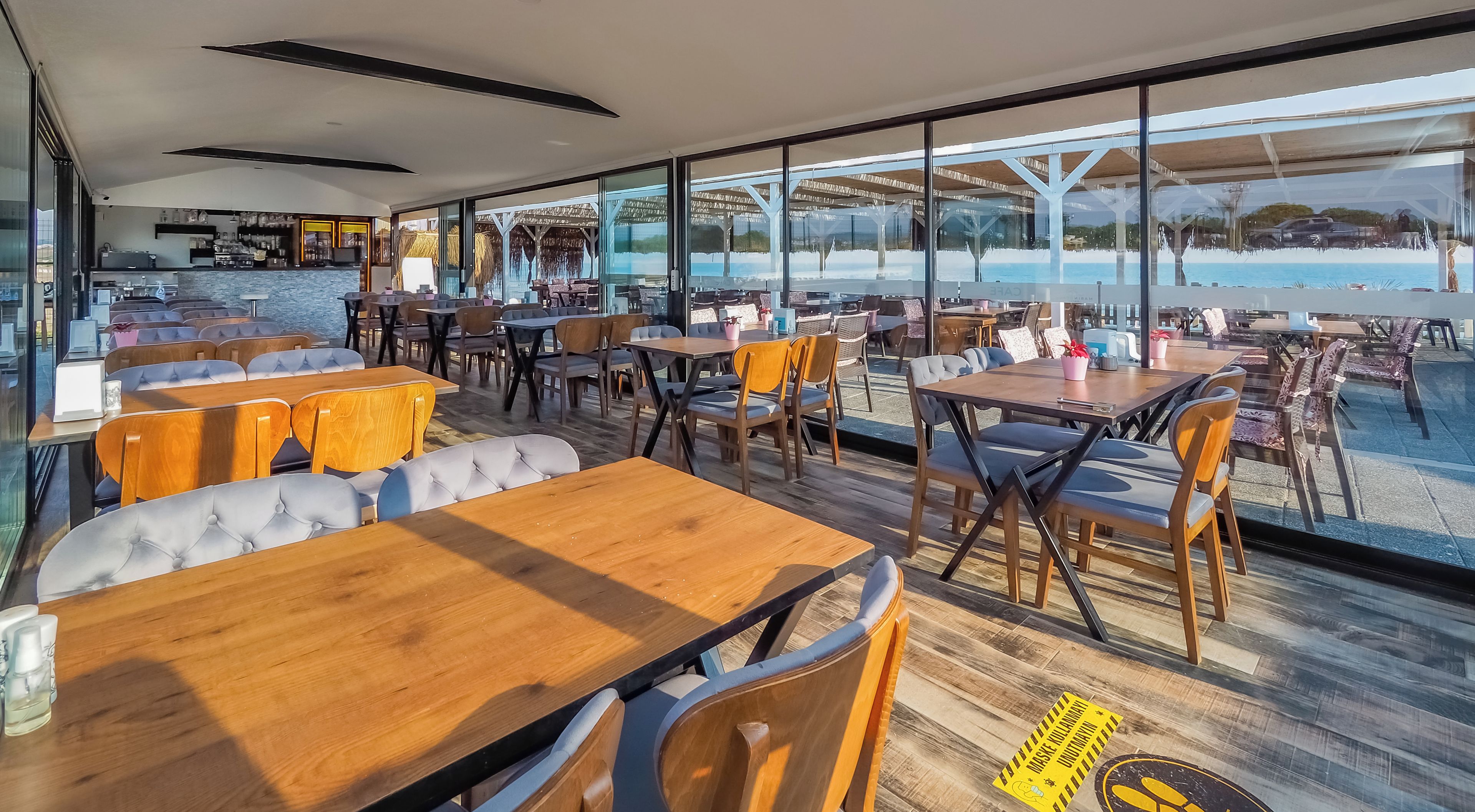 Un restaurante con mesas y sillas de madera, ventanas amplias y vista al mar.