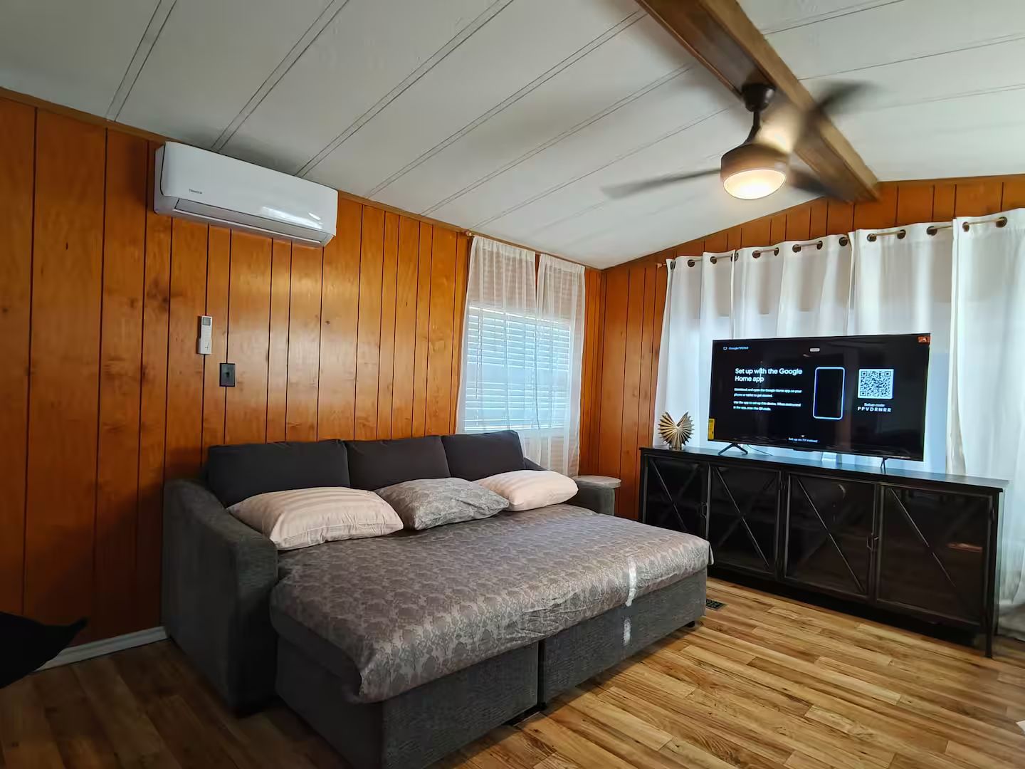 Une chambre avec un lit, des murs en bois, un ventilateur de plafond, un appareil de climatisation, une télévision et une fenêtre avec des rideaux.