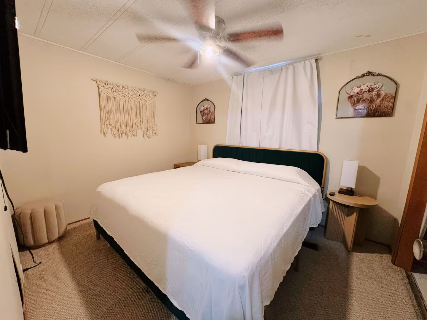 Una habitación de hotel con cama, ventilador de techo, mesita de noche con lámpara, una obra de arte en la pared y una ventana con cortinas.