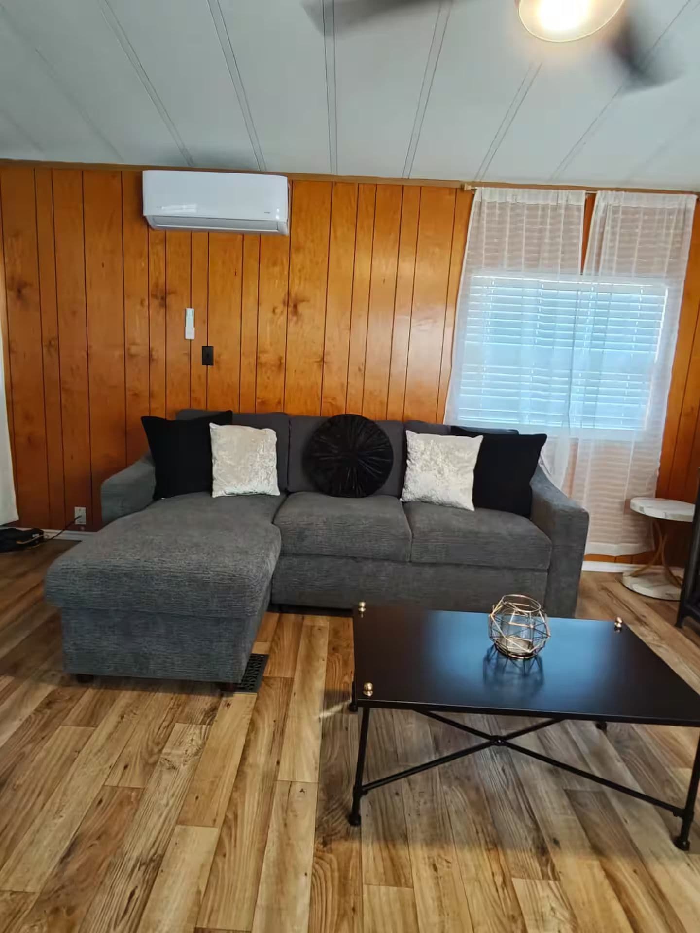 Una sala de estar con un sofá seccional gris, una mesa de centro negra y piso de madera.