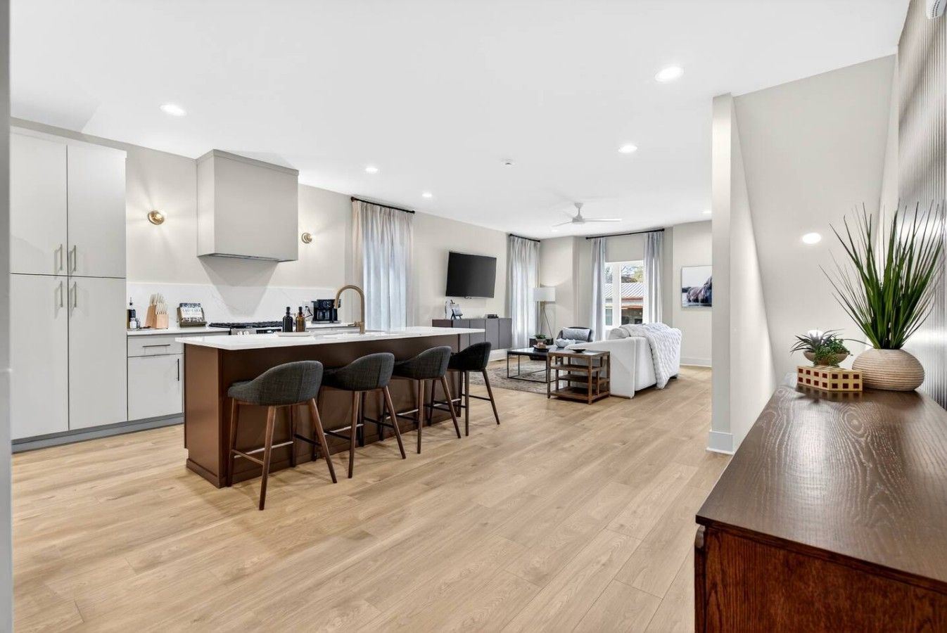 A modern kitchen with a central island, bar stools, a dining area with a table and chairs, and a living room with a sofa and a TV.