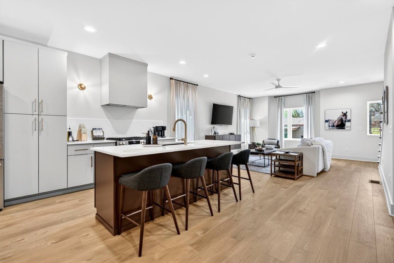 A modern kitchen with a central island, bar stools, a dining area with a table and chairs, and a living room with a sofa and a TV.
