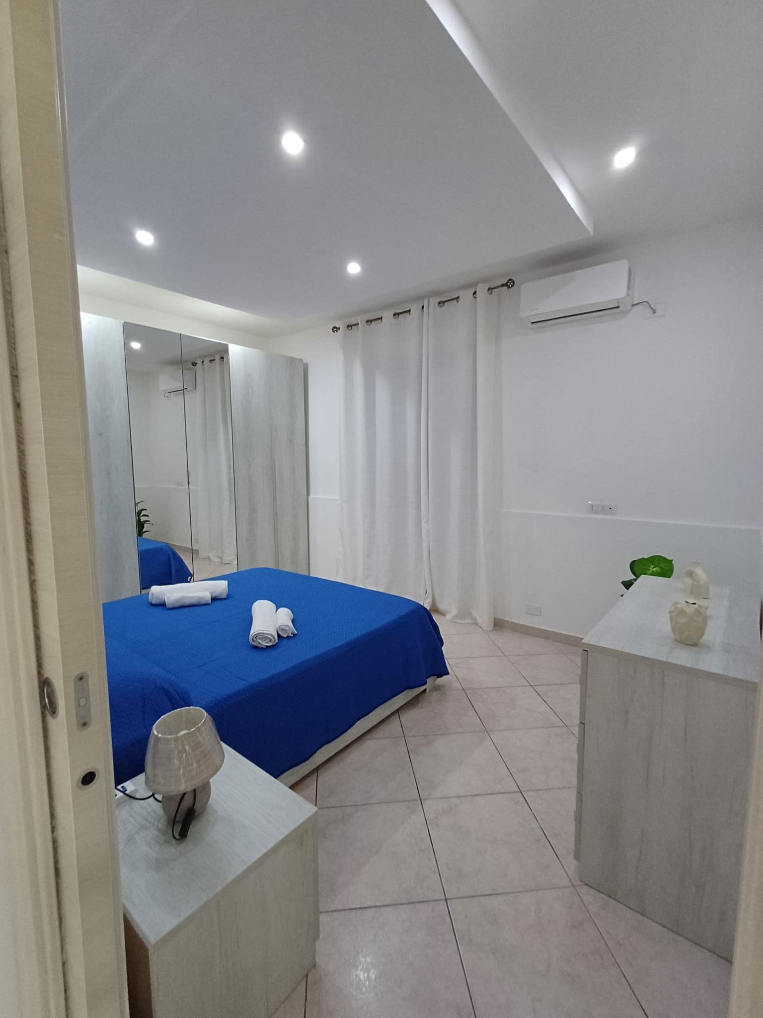 A hotel room with a bed, a mirror, a small table with a lamp, and a bathroom area with a sink and a green plant.