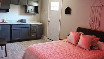 A bedroom with a large bed, a nightstand, a dresser, and a kitchen area with cabinets and a countertop.