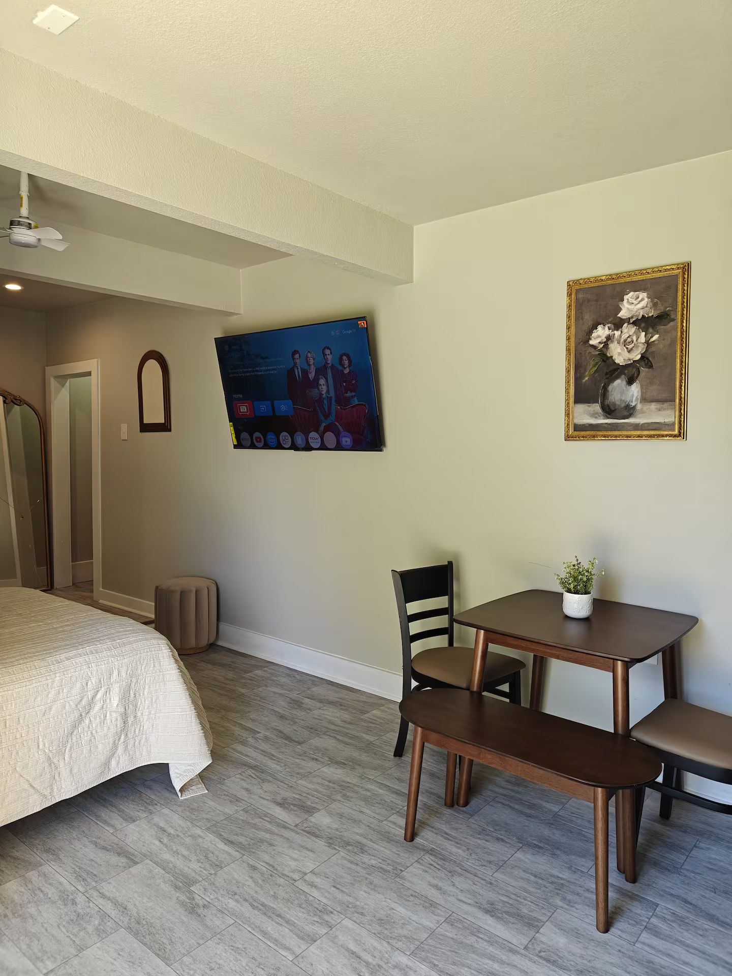 Une chambre d’hôtel avec un lit, un téléviseur fixé au mur, un tableau, une petite table avec un banc et une plante en pot.