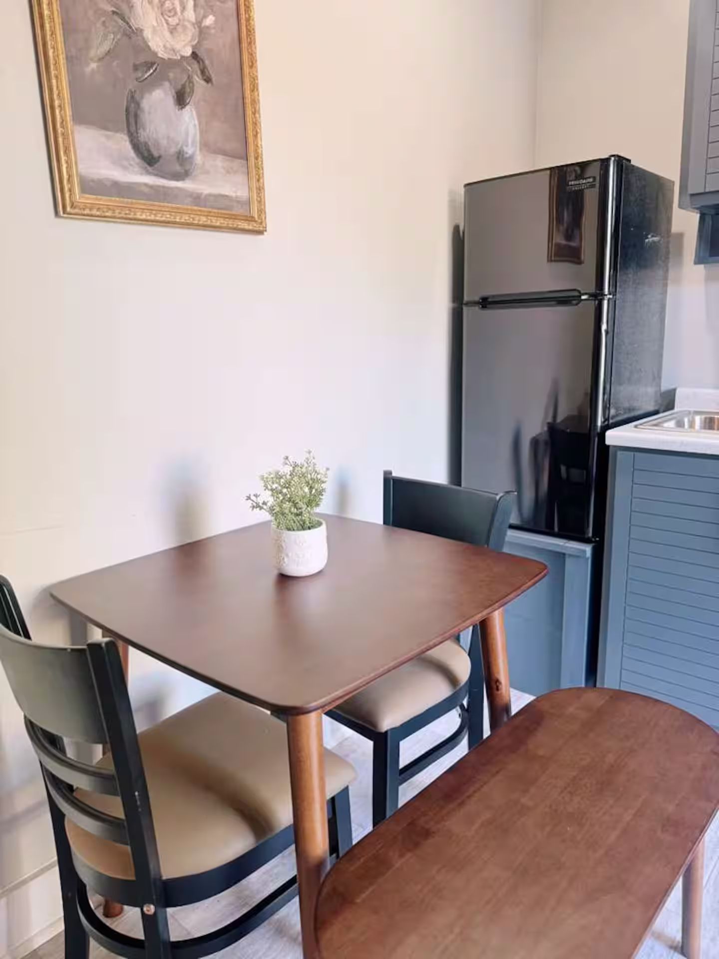 A small dining area with a table and chairs, a potted plant, and a refrigerator in the background.