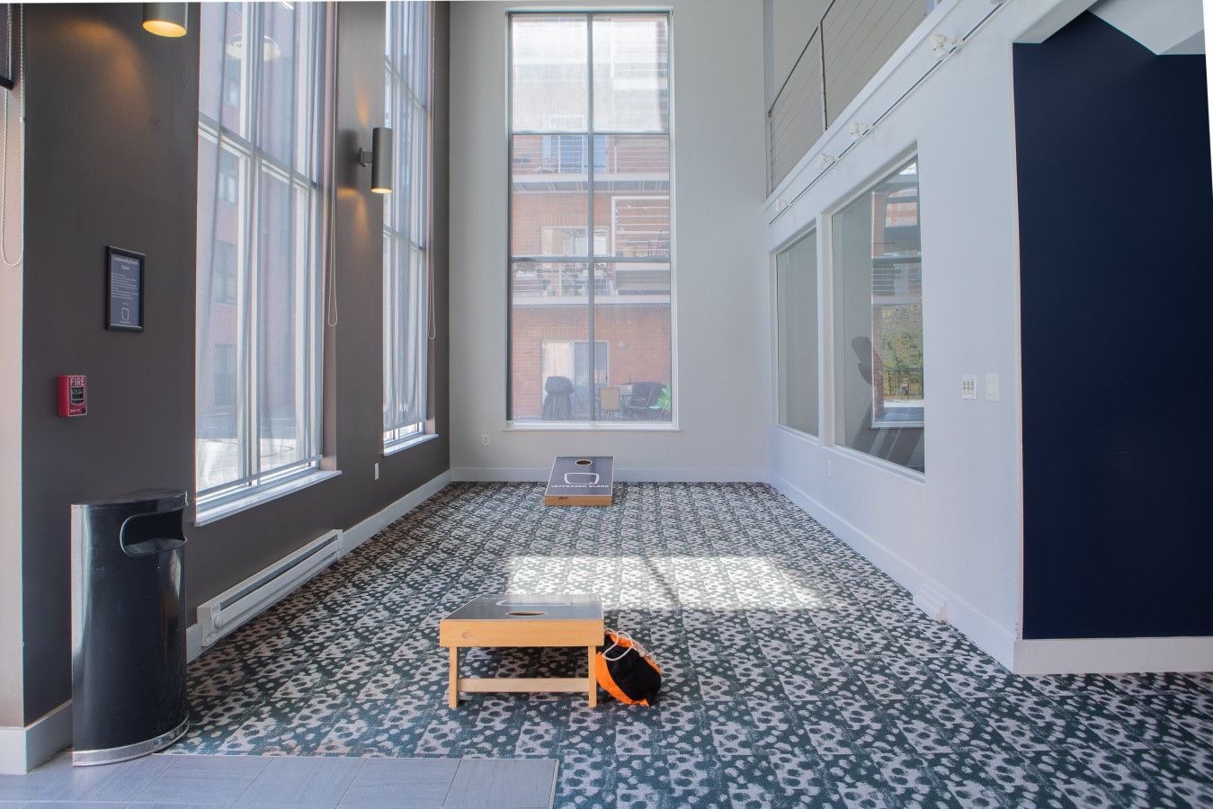 A room with large windows, a fire alarm, a trash bin, and a small wooden table.