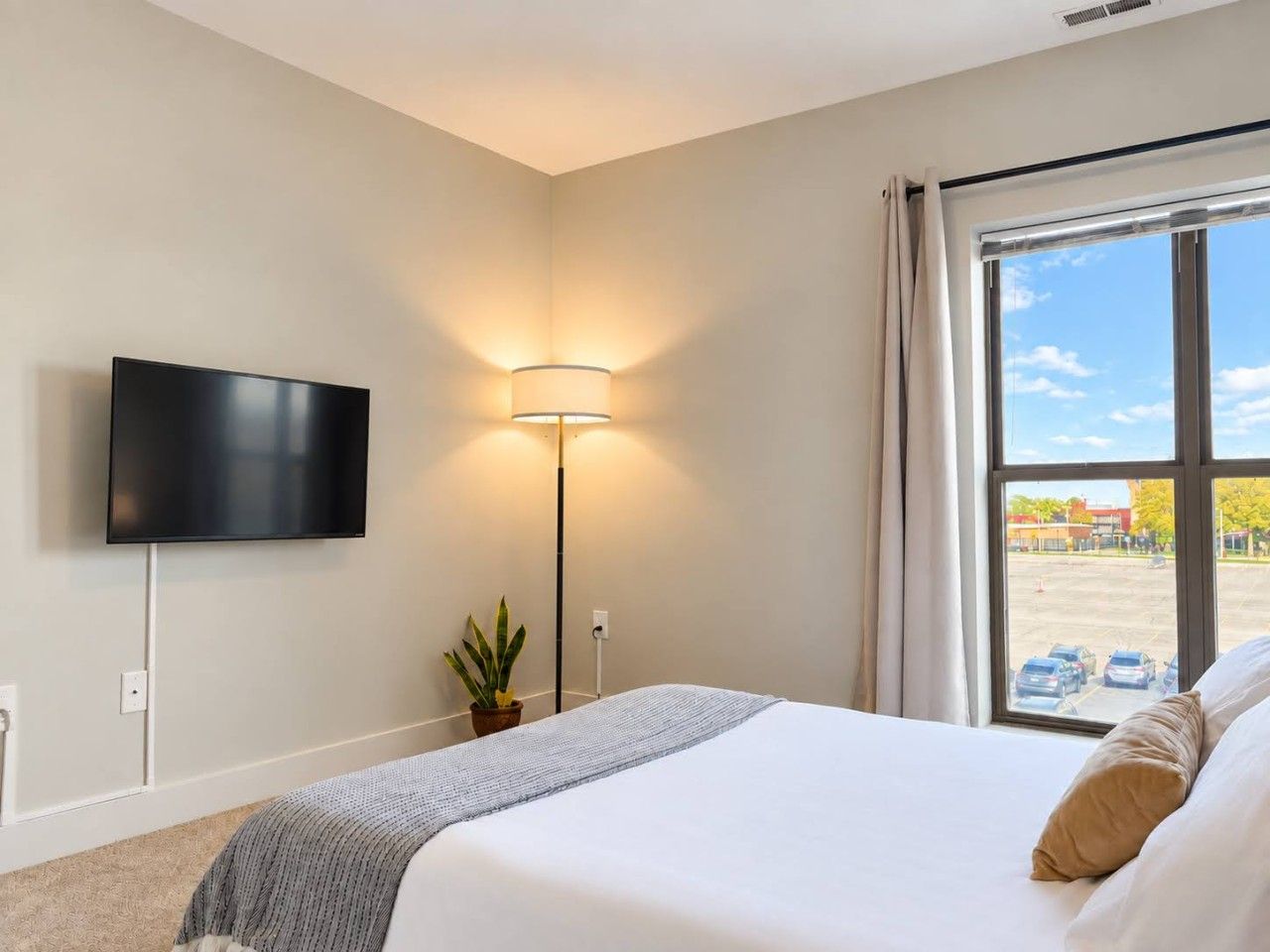 A hotel room with a bed, a flat-screen TV mounted on the wall, a floor lamp, a window with a view of a parking lot, and a potted plant.