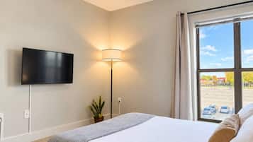 A hotel room with a bed, a flat-screen TV mounted on the wall, a floor lamp, a window with a view of a parking lot, and a potted plant.