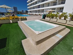 Una piscina con idromassaggio, circondata da erba sintetica e piante in vaso, con un edificio moderno sullo sfondo.