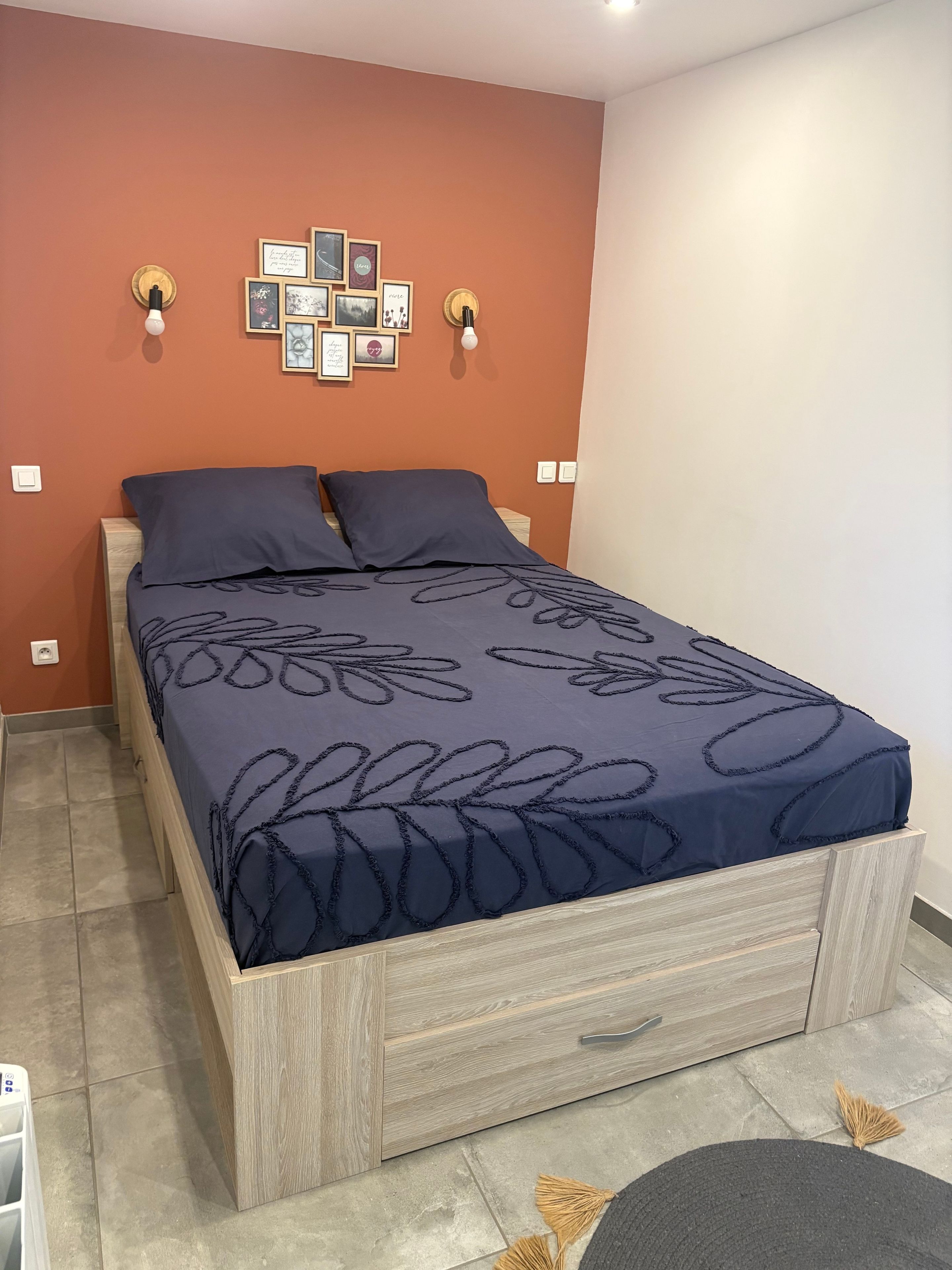 A bedroom with a bed, a wooden nightstand, a wall-mounted light fixture, and a wall decoration.