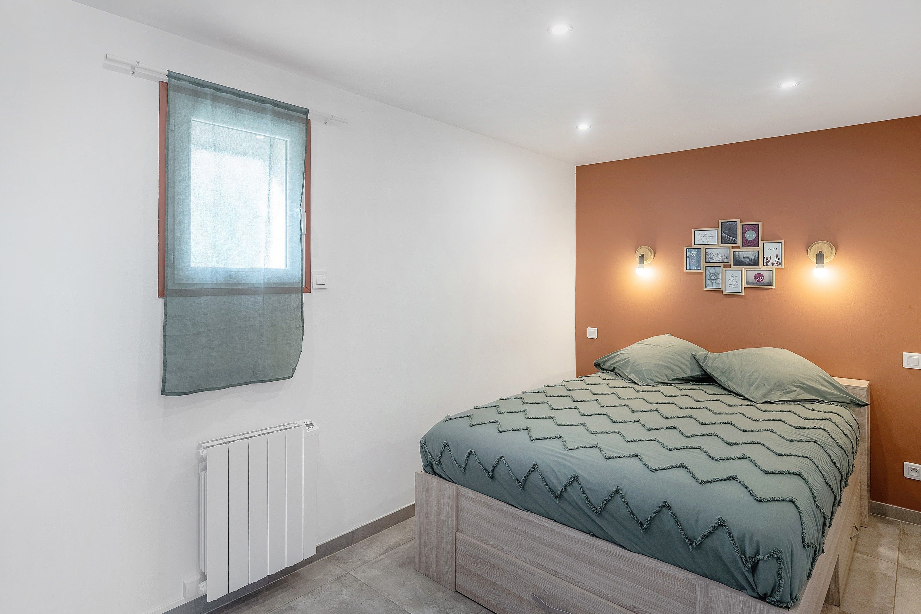 A bedroom with a bed, a radiator, a window with a blind, and wall-mounted picture frames.