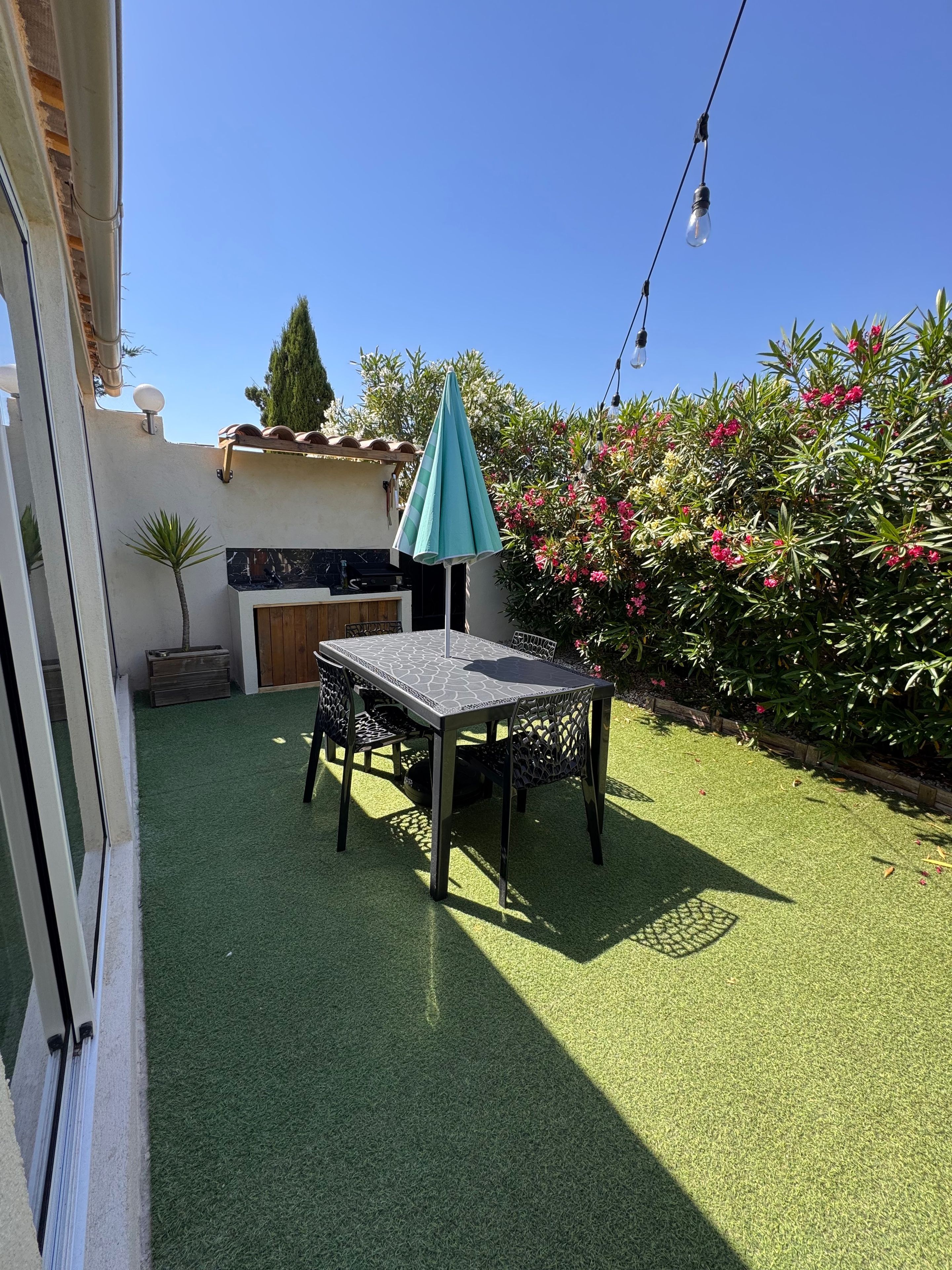 A patio with artificial grass, a table and chairs, a teal umbrella, and a string of lights.