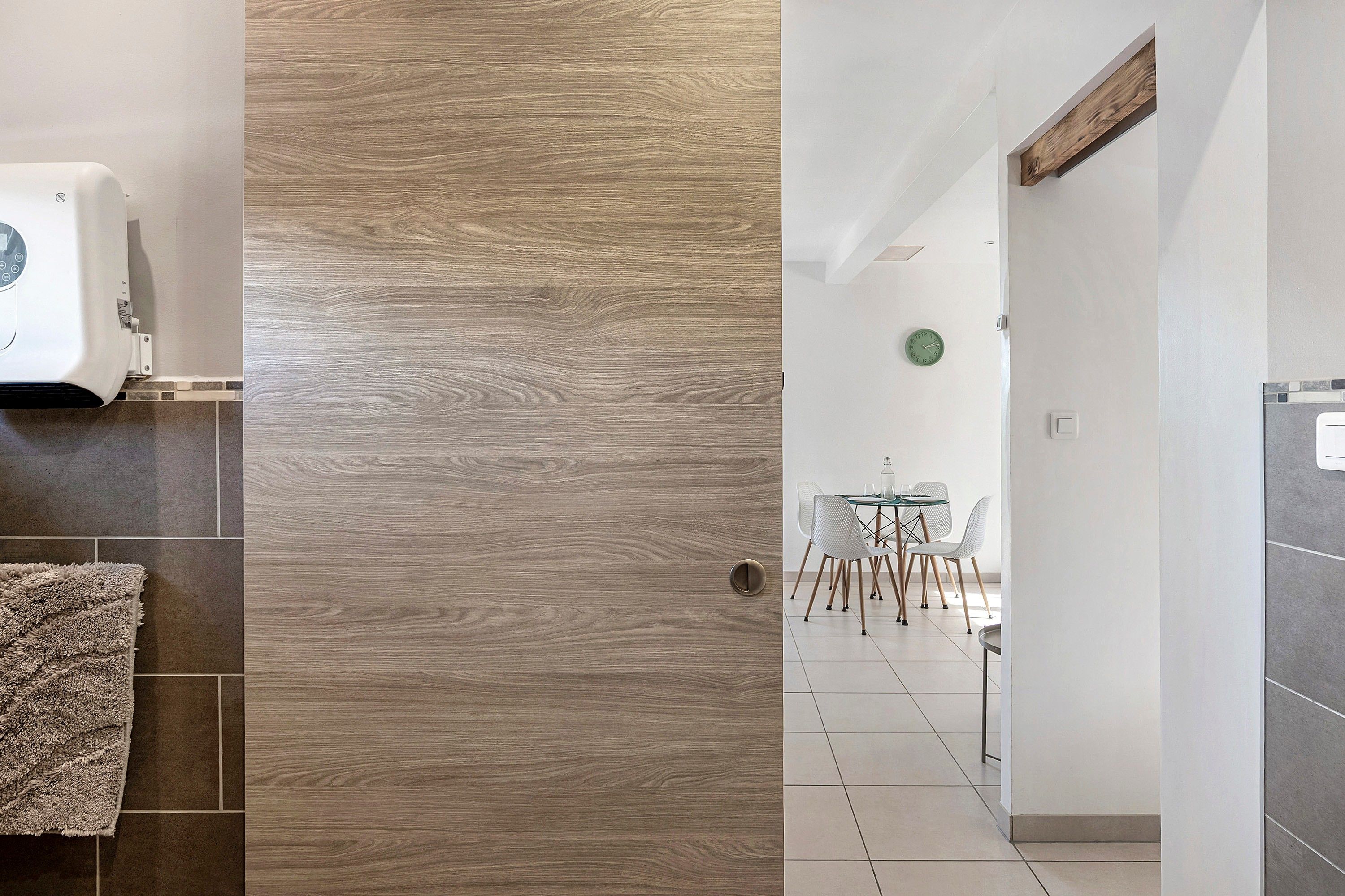 Un pasillo con pared de madera, suelo de baldosas, pared blanca con un reloj y un comedor con sillas y una mesa.
