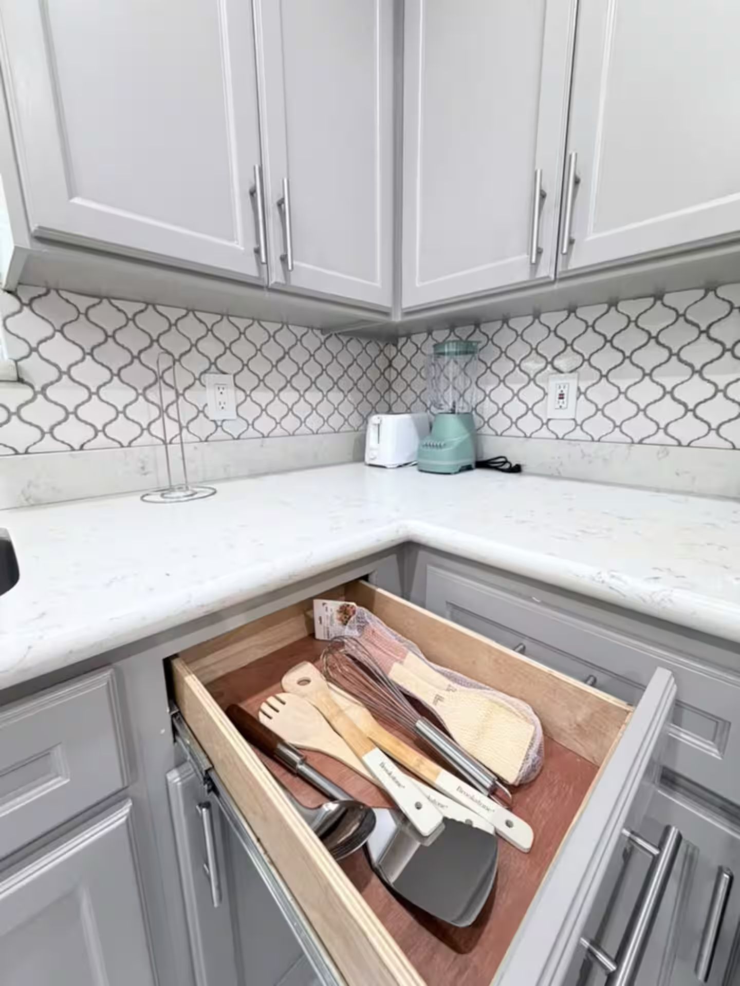A kitchen with white cabinets, a patterned backsplash, and a drawer containing wooden utensils.