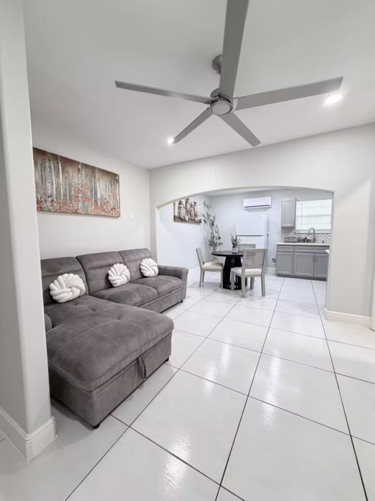 A modern living room with a grey sofa, a ceiling fan, and a kitchen in the background.
