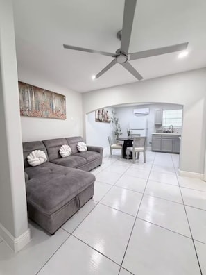 A modern living room with a grey sofa, a ceiling fan, and a kitchen in the background. - 1717 Apartment (Laredo)