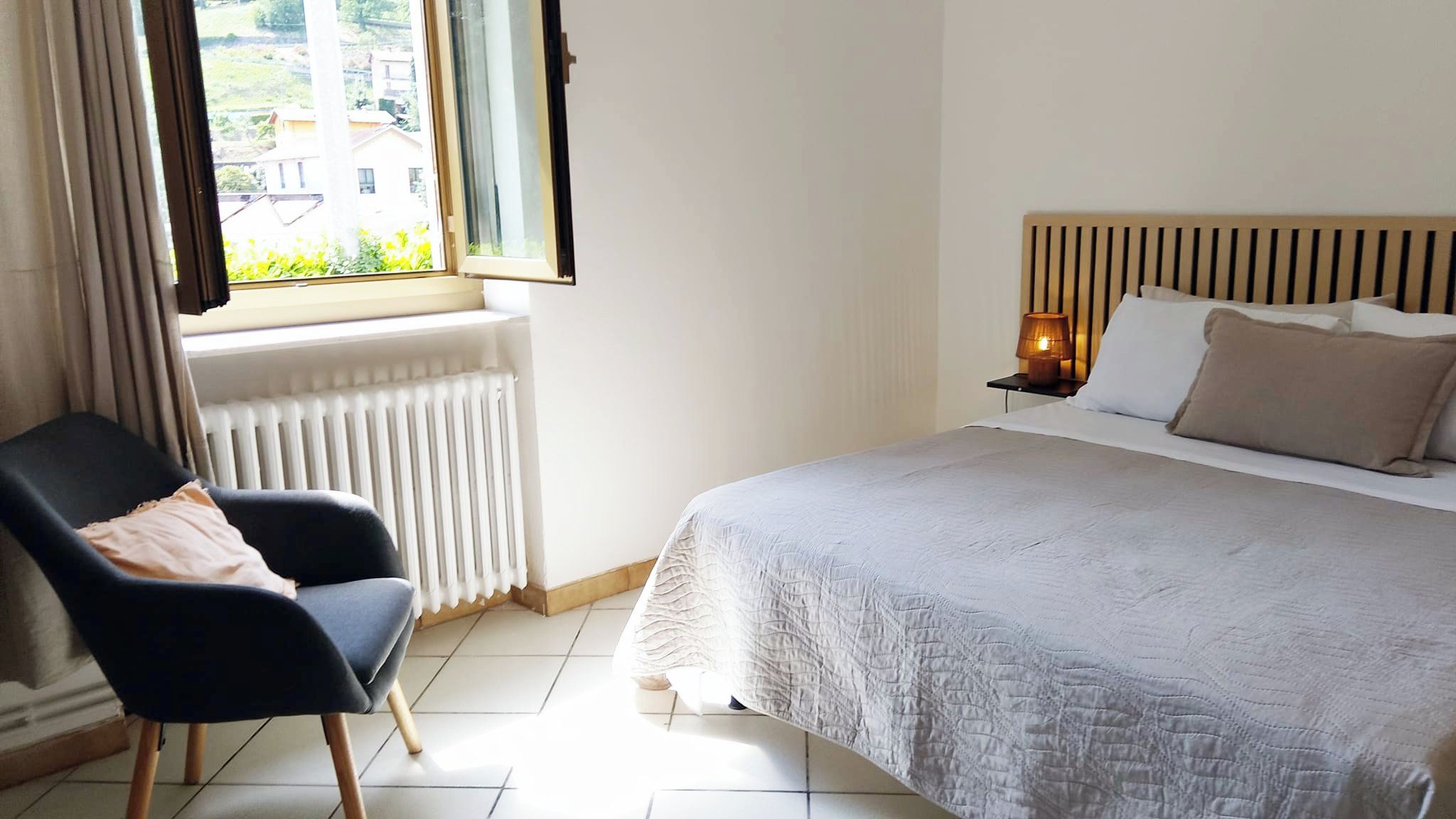 A bedroom with a bed, a chair, a window, a radiator, and a nightstand with a lamp.
