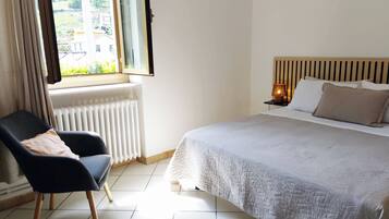 A bedroom with a bed, a chair, a window, a radiator, and a nightstand with a lamp.