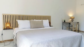 Una cama bien tendida con cabecera, una lámpara de noche y una mesita con lámpara y planta.