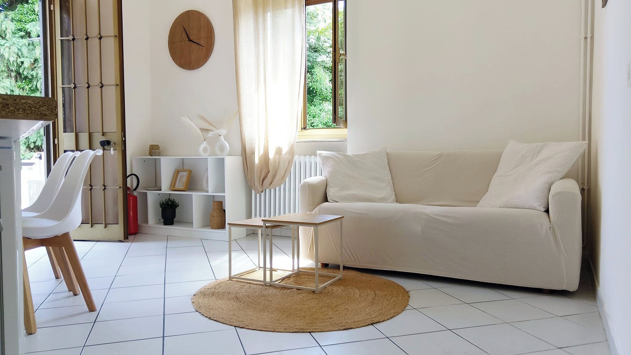 A modern living room with a white sofa, a small wooden table, and a white chair. There is a clock on the wall and a window with curtains.