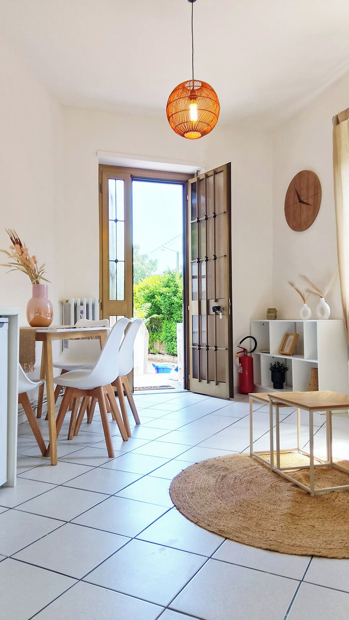 A modern living room with a dining area, featuring a round table, white chairs, a woven floor lamp, and a large window with a view of greenery.