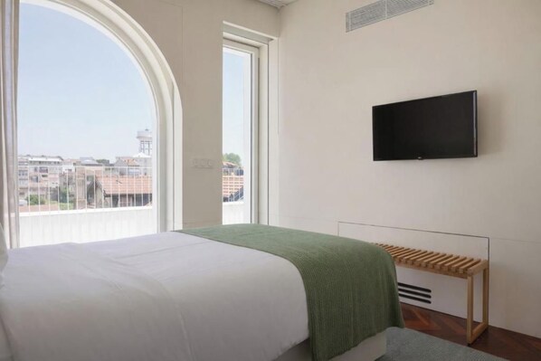 Ein Hotelzimmer mit Bett, einem an der Wand befestigten Fernseher und einem Fenster mit Blick auf Gebäude.