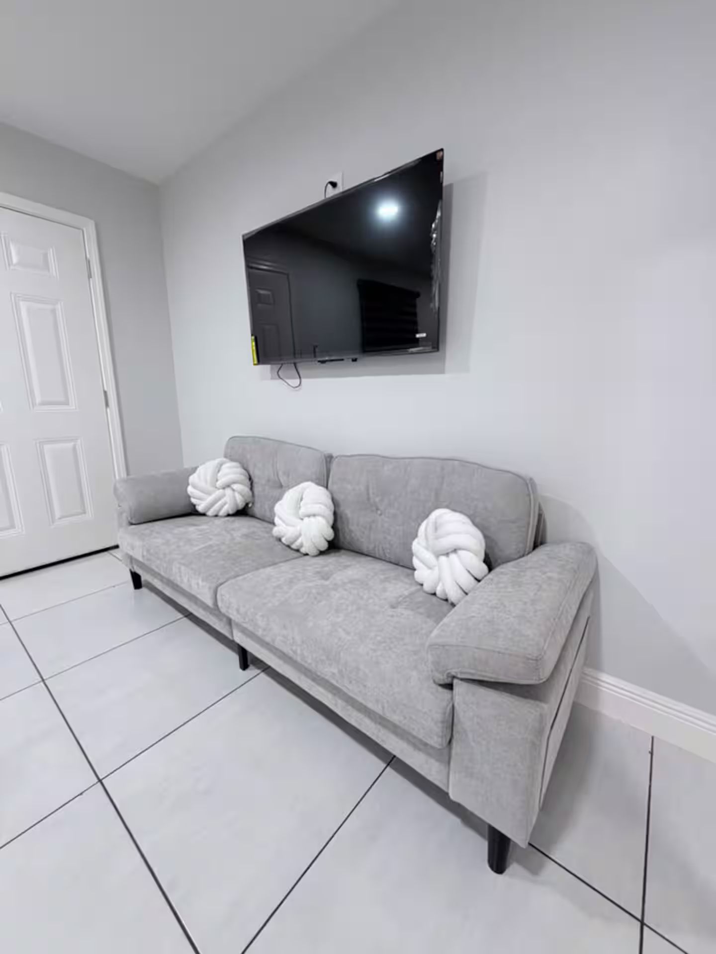 A grey sofa with three white, twisted pillows against a white wall, a flat-screen TV mounted above it, and a white door to the left.