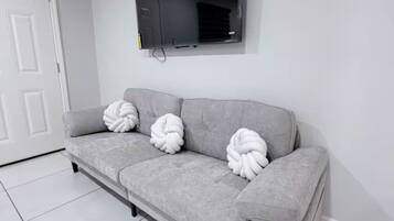 A grey sofa with three white, twisted pillows against a white wall, a flat-screen TV mounted above it, and a white door to the left.