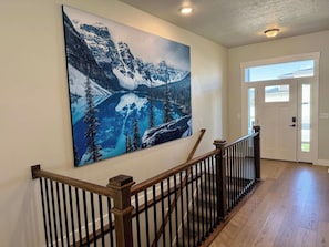 A hallway with a wooden railing, a large mountain landscape painting on the wall, and a door with a view of the outside. - Heber Valley Escape | Swim Spa + RV Pad, Sleeps 11 (Heber City)