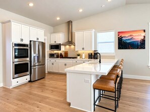 A modern kitchen with white cabinetry, stainless steel appliances, a central island with bar stools, and a large window with a mountain view. - Heber Valley Escape | Swim Spa + RV Pad, Sleeps 11 (Heber City)