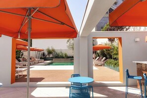 Un espace piscine extérieur agrémenté de parasols orange, de chaises longues et d’un bar avec des chaises bleues.