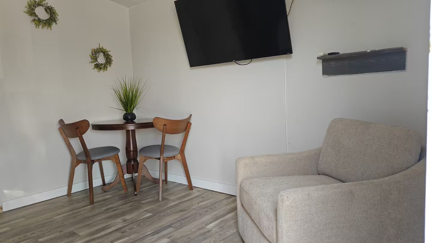 Una habitación con un sofá gris, una mesa de comedor de madera con dos sillas, una planta en maceta y un televisor de pantalla plana montado en la pared.