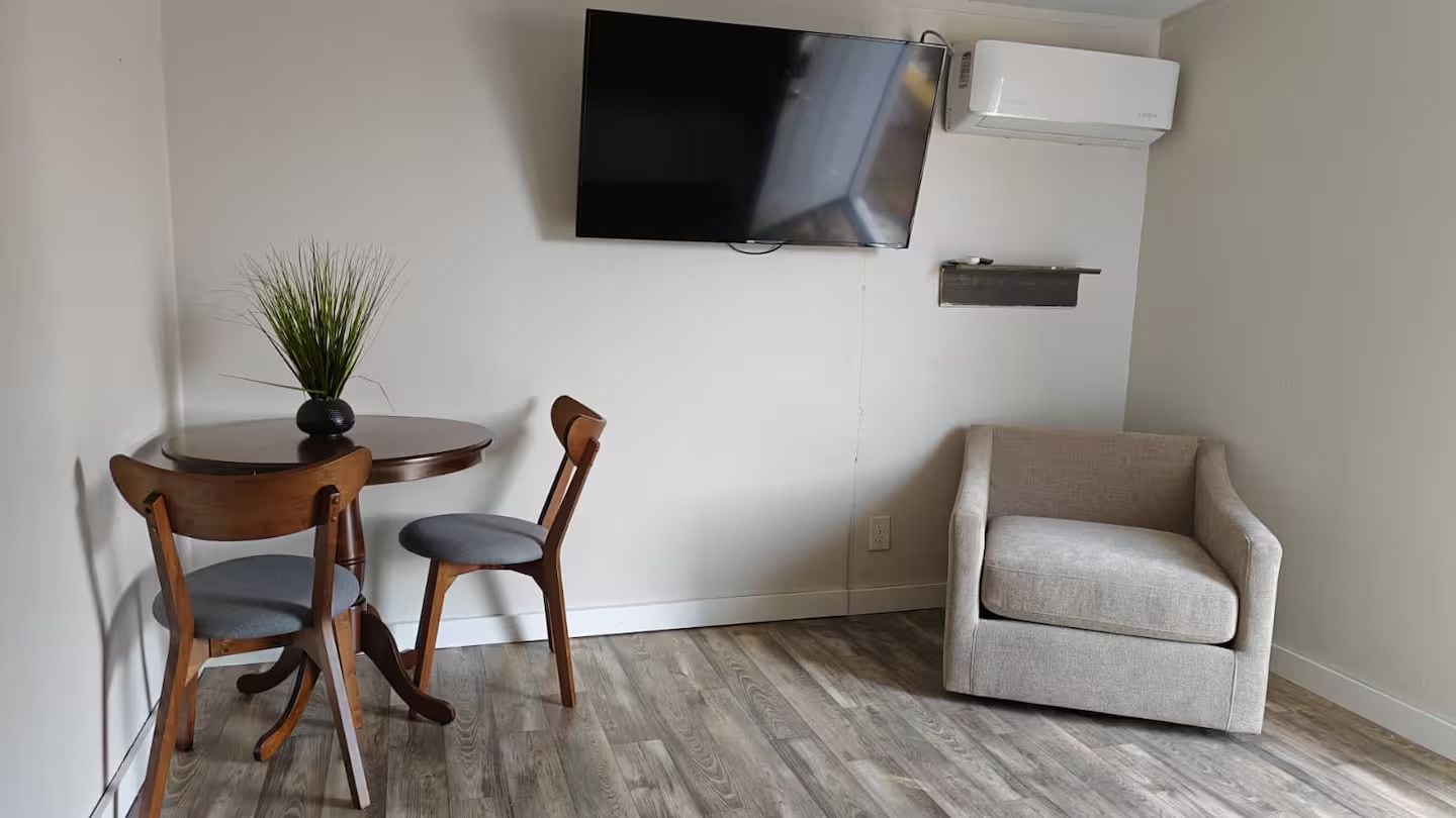 Una habitación con una televisión de pantalla plana montada en la pared, una mesita redonda con dos sillas, una planta en maceta y un sofá color beige.