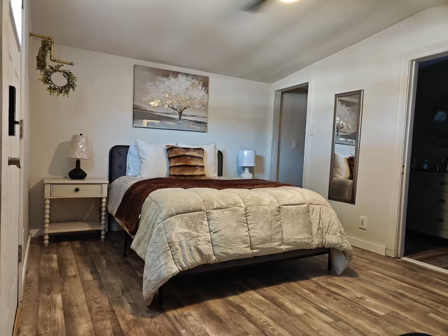 Un dormitorio con una cama, dos mesitas de noche, una lámpara de pared, un cuadro y una puerta que da a otra habitación.