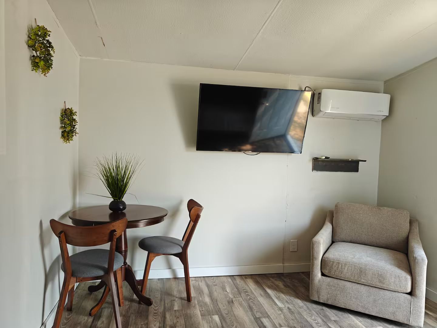 Una sala de estar con un televisor de pantalla plana montado en la pared, una mesa redonda de madera con dos sillas, un sillón beige y una pequeña planta en maceta sobre la mesa.