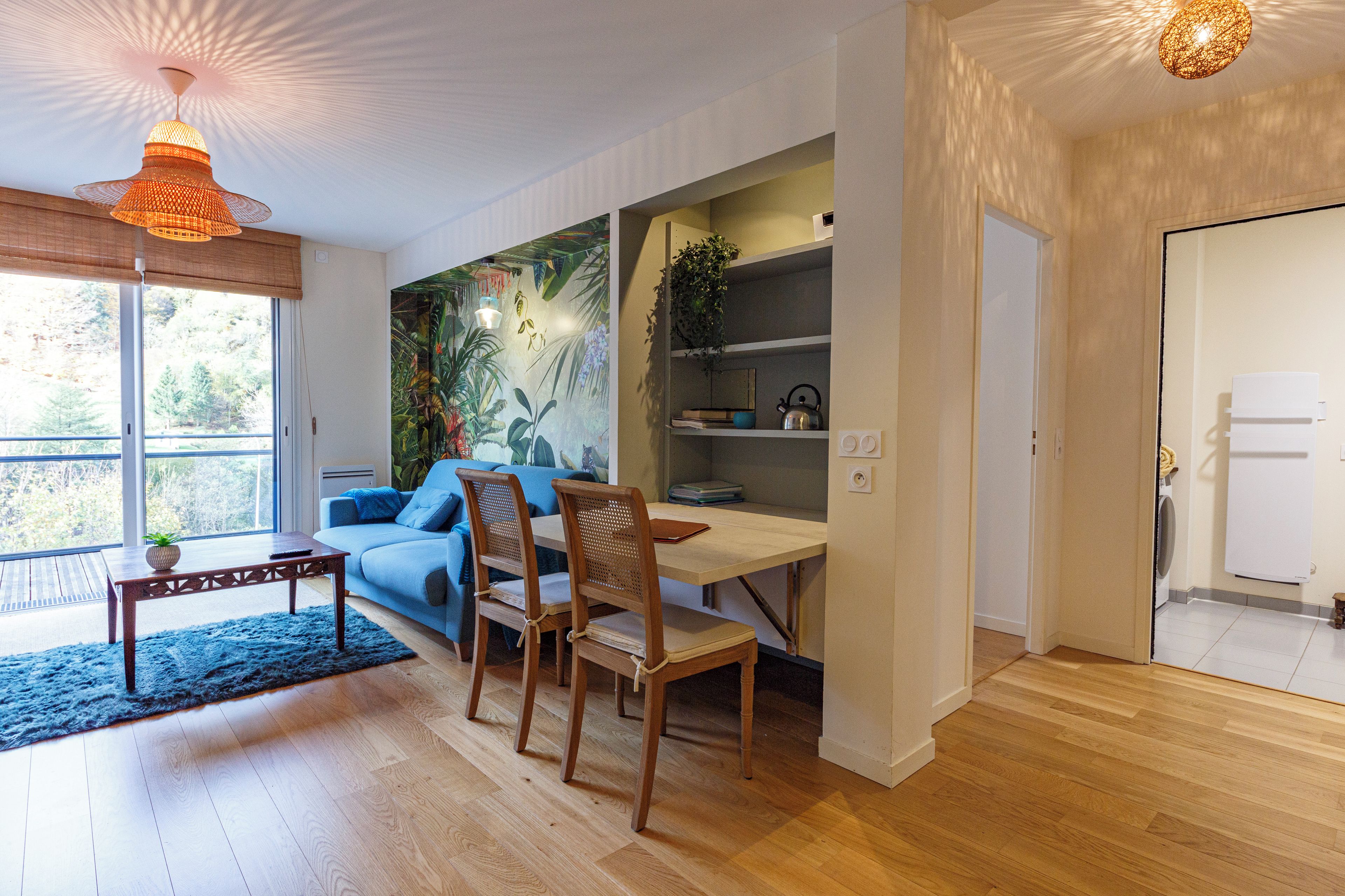 Ein modernes Wohnzimmer mit einem blauen Sofa, einem Holzespeisetisch und Stühlen. Ein großes Fenster lässt natürliches Licht herein und bietet einen Ausblick ins Freie.