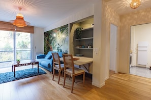 A modern living room with a blue sofa, wooden dining table, and chairs. There is a large window providing natural light and a view of the outdoors. - Le B3 de Claire (Mont-Dore)