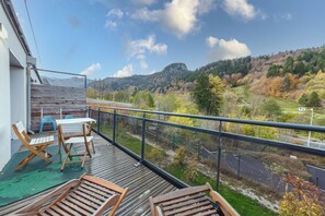 A balcony with a view of a mountainous landscape, a wooden deck, and a table with chairs. - Le B3 de Claire (Mont-Dore)