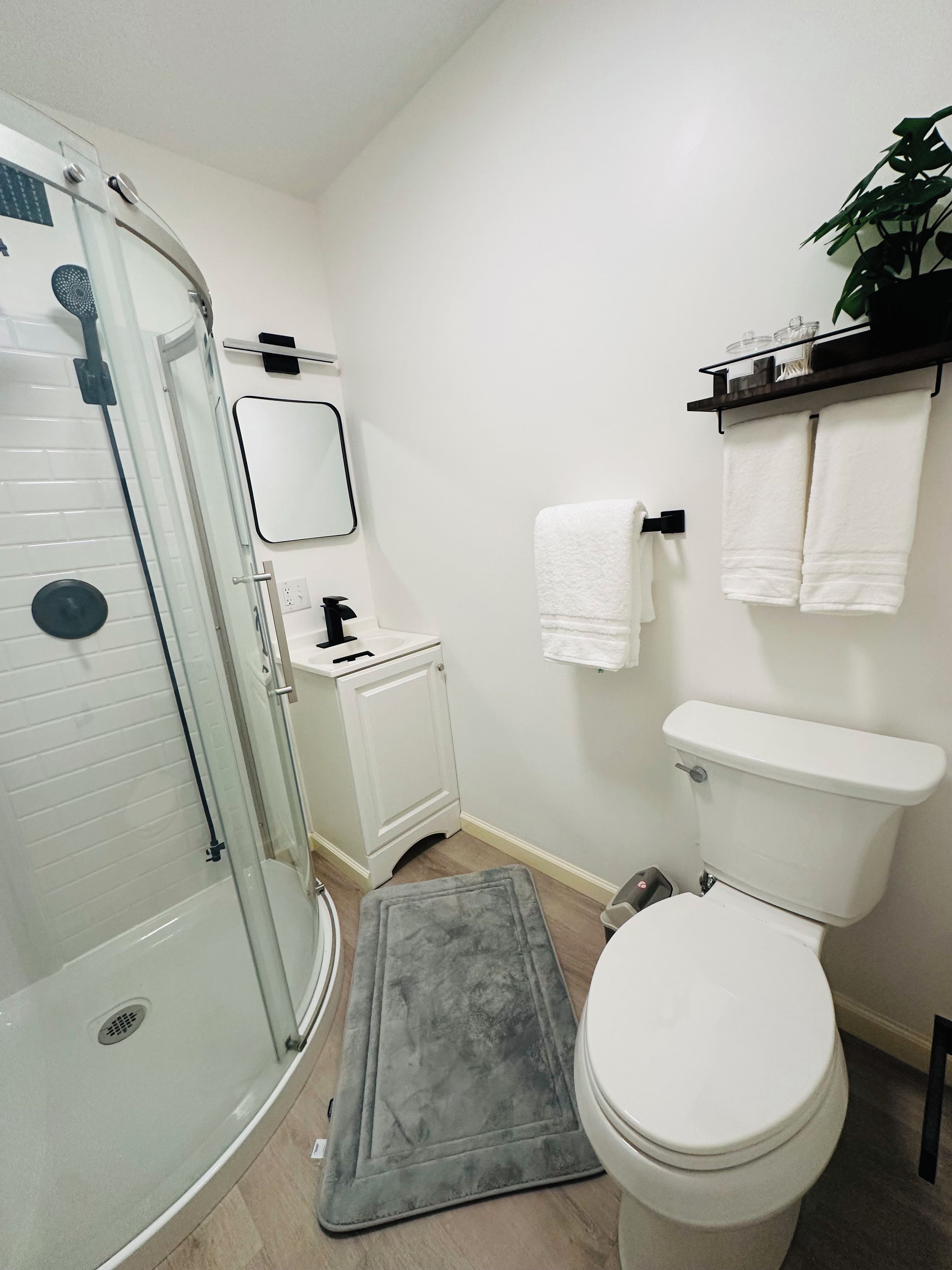 A bathroom with a glass shower enclosure, a white toilet, a sink with a mirror, and a shelf with towels.