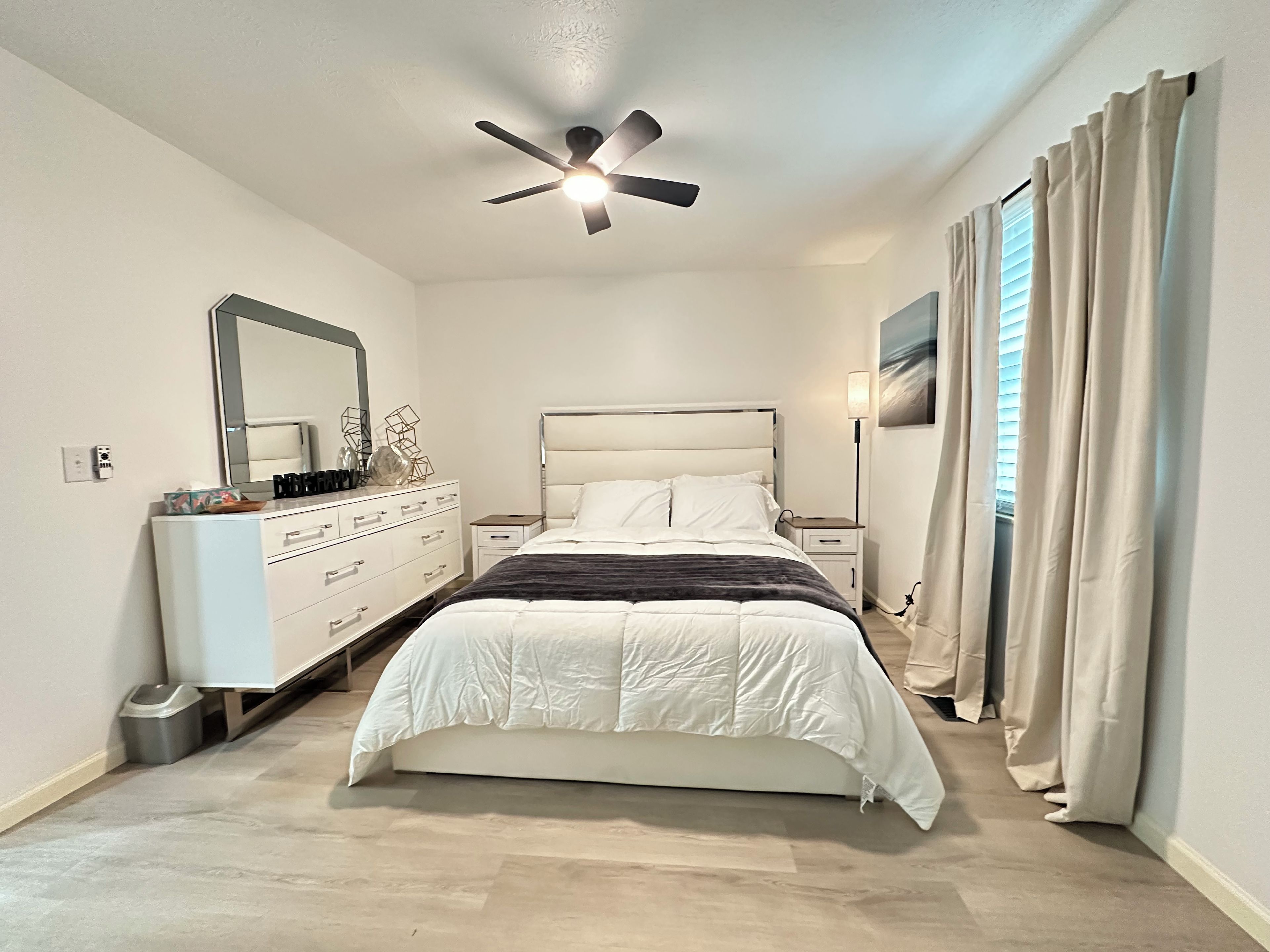 A bedroom with a bed, dresser, ceiling fan, and curtains.