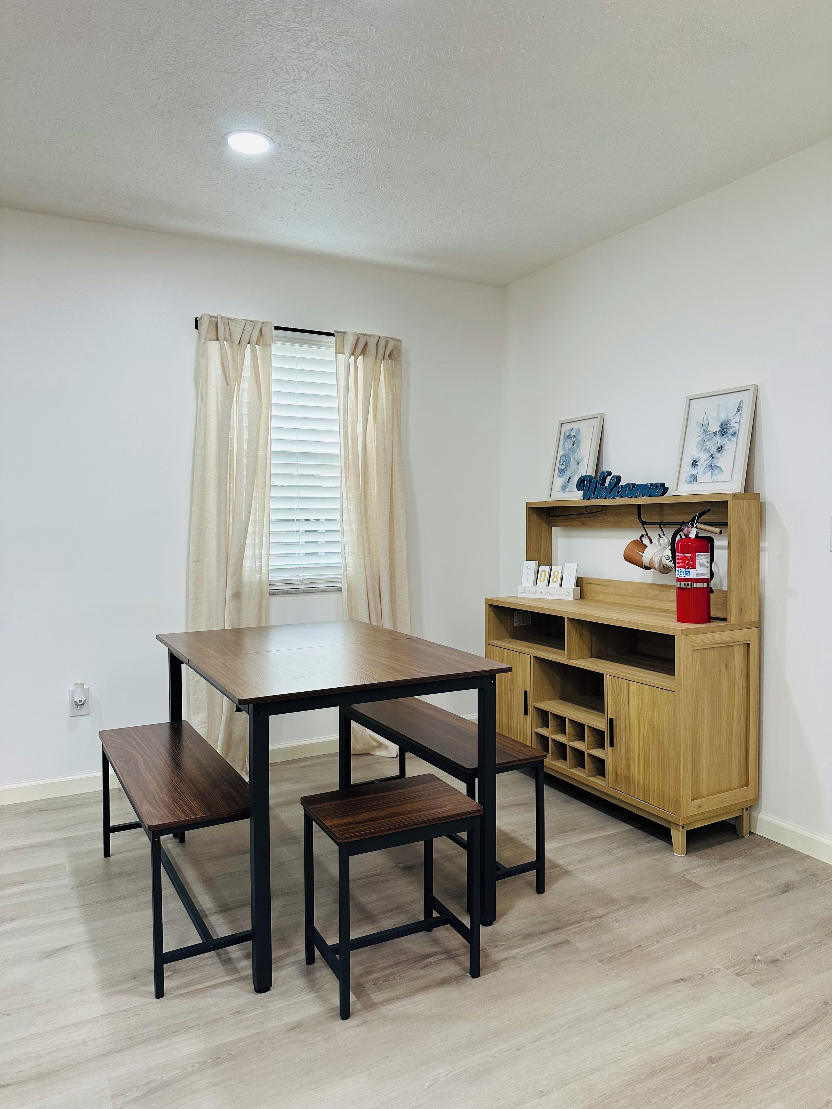 A room with a wooden table and benches, a wooden cabinet with a red fire extinguisher, and a window with curtains.