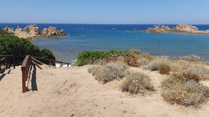 A sandy beach with a wooden walkway leading to the sea, surrounded by rocky formations and sparse vegetation. - Beach Home in Sardegna Italy - Wake Up in Heaven! (Canneddi)
