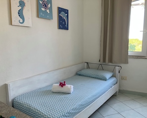 A single bed with patterned bedding, a wall-mounted light switch, and a window with curtains. - Beach Home in Sardegna Italy - Wake Up in Heaven! (Canneddi)