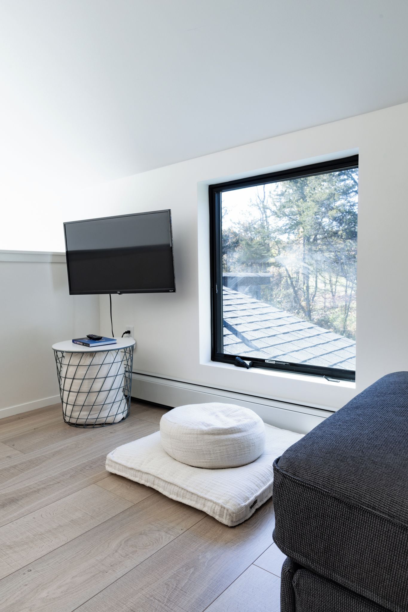 A room with a flat-screen TV mounted on the wall, a round white ottoman, a small round table with a book and remote, and a large window with a view of trees and a roof.